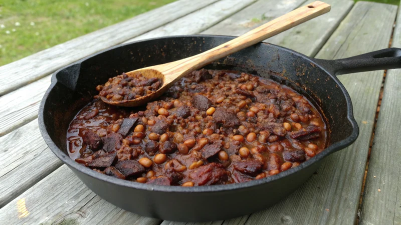 Close-up of pinto beans in dark molasses-ancho sauce with chopped brisket, glossy mahogany color, cast iron rim visible