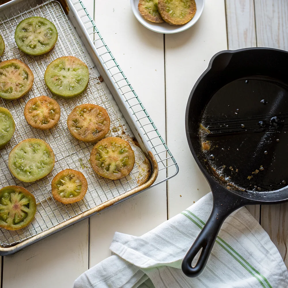 Overhead view of fried green tomato slices arranged on a wire rack over a sheet pan next to a black cast iron skillet with shimmering oil on a wooden counter