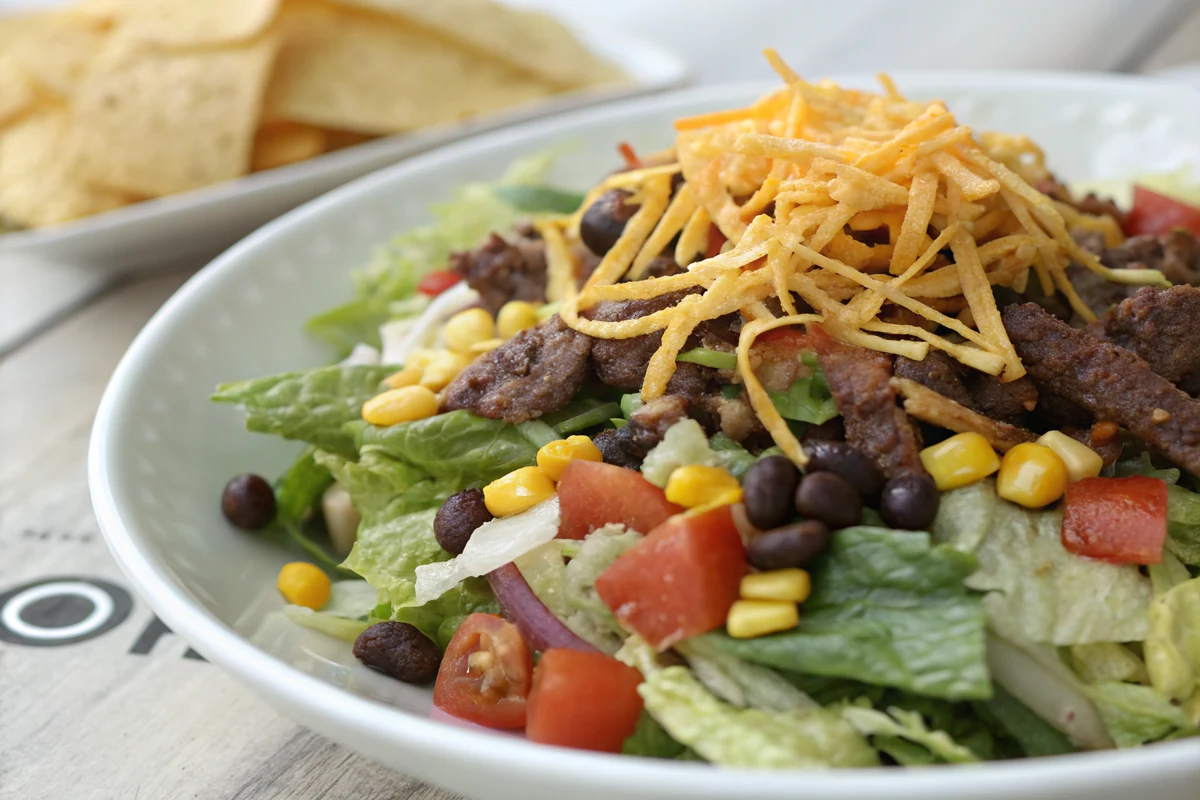 Close-up of a Tex-Mex salad showing layers of crisp romaine, spiced beef, black beans, corn, diced tomato, shredded cheddar, and fried tortilla strips