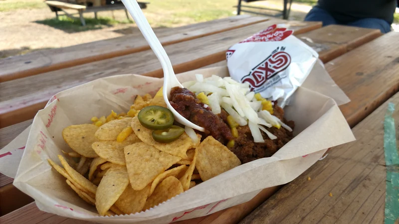 Close-up of a fork lifting Fritos coated in chili and melted cheddar from a slit-open bag, jalapeno slice visible on top