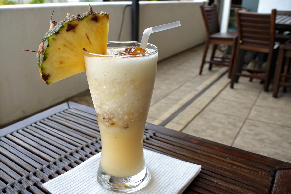 Tall glass of thick frozen pina colada with a thin layer of dark rum floating on the surface and a notched pineapple wedge on the rim