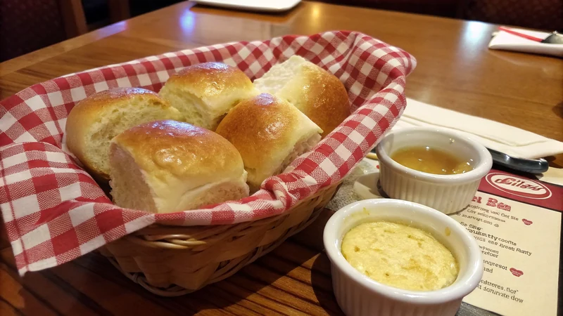 Close-up of a torn-open Texas Roadhouse roll showing pillowy white crumb and a knife spreading cinnamon honey butter into the steam